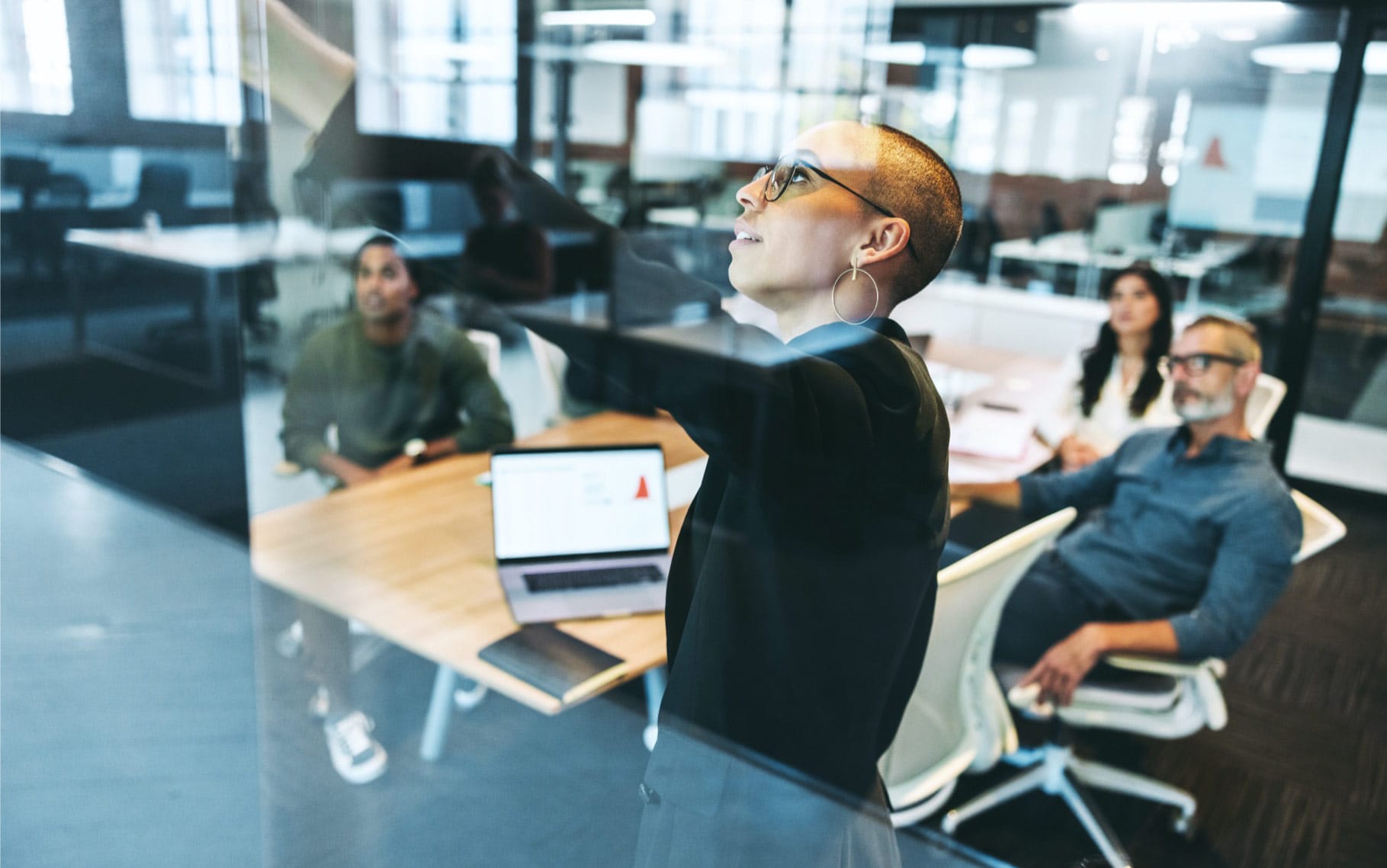 Person with short hair and glasses presenting in a modern office meeting with colleagues seated at a table with laptops.