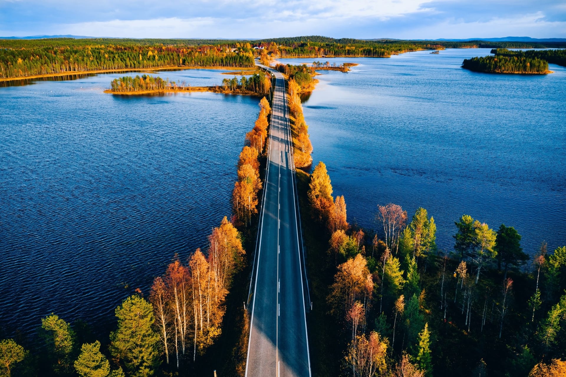 Aerial view of a straight road crossing blue lakes, lined with autumn trees in golden and orange hues.