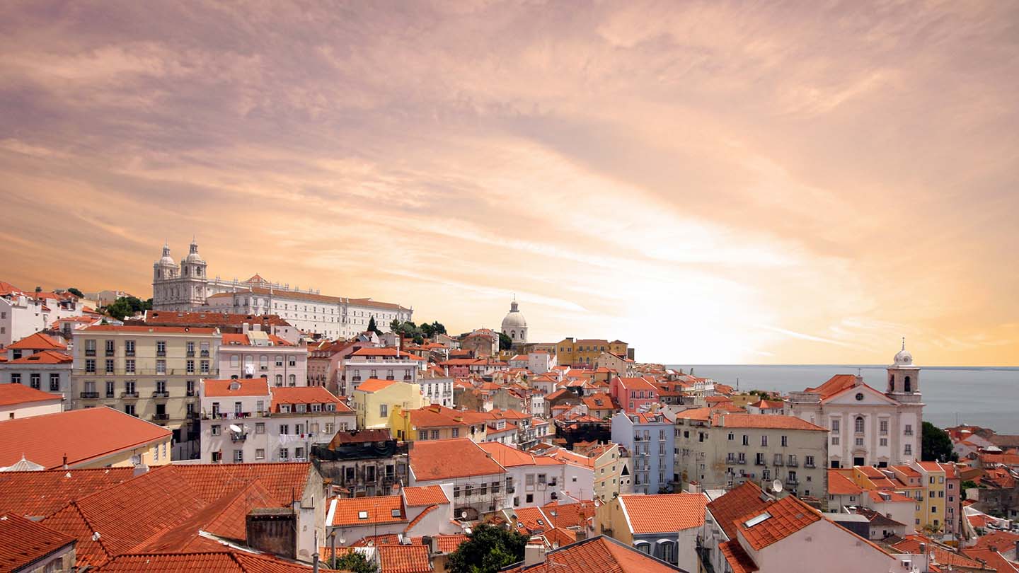 Lisbon cityscape with orange terracotta rooftops, historic buildings and cathedral under a golden sunset sky.