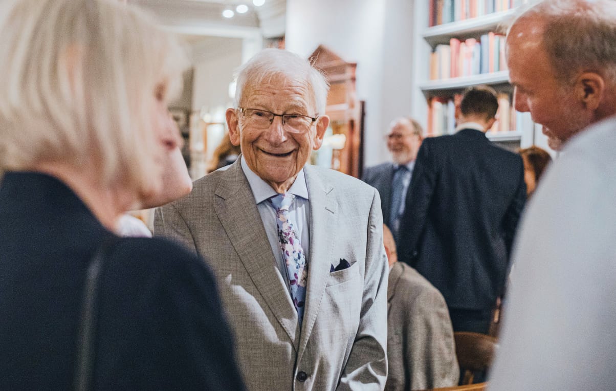 Elderly gentleman in light grey suit with floral tie smiling at social gathering in a library setting.