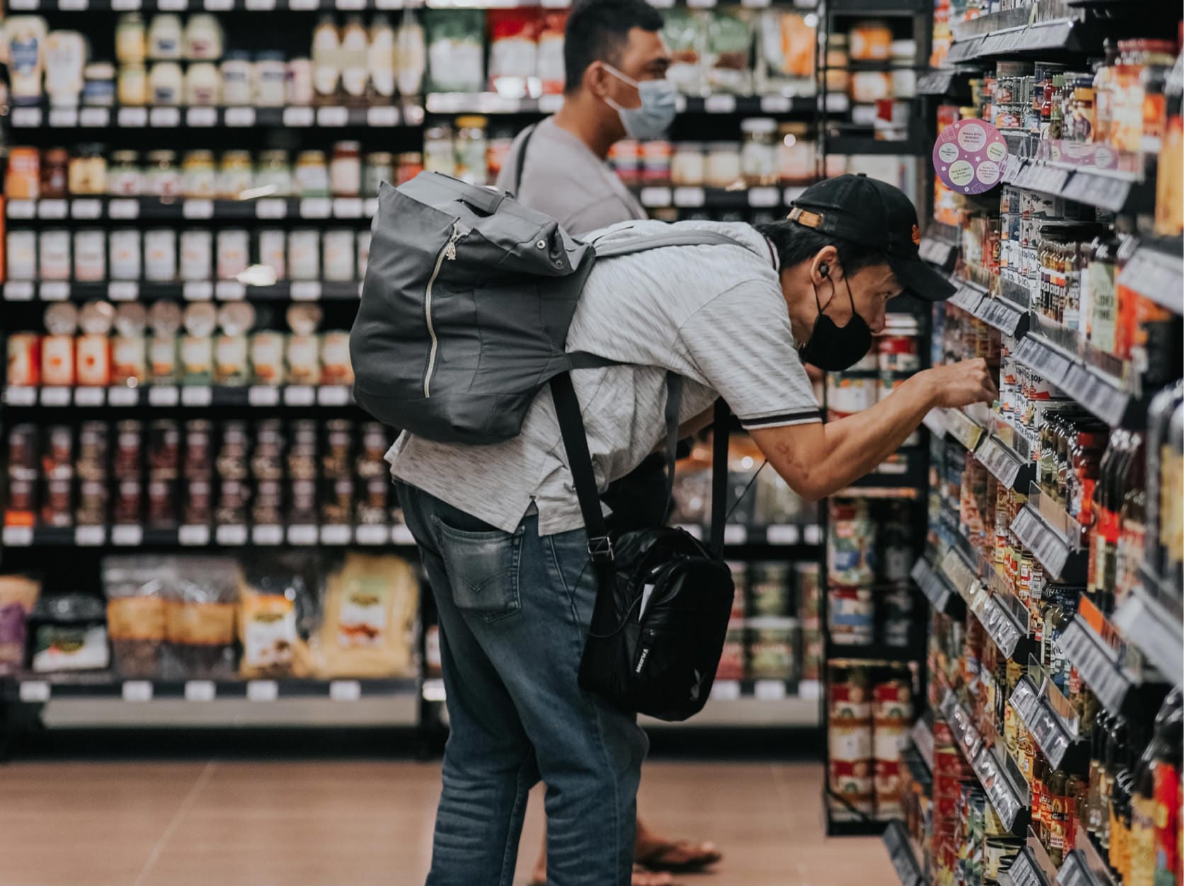 Shoppers wearing face masks browse grocery store shelves, one with a grey backpack examining products closely.