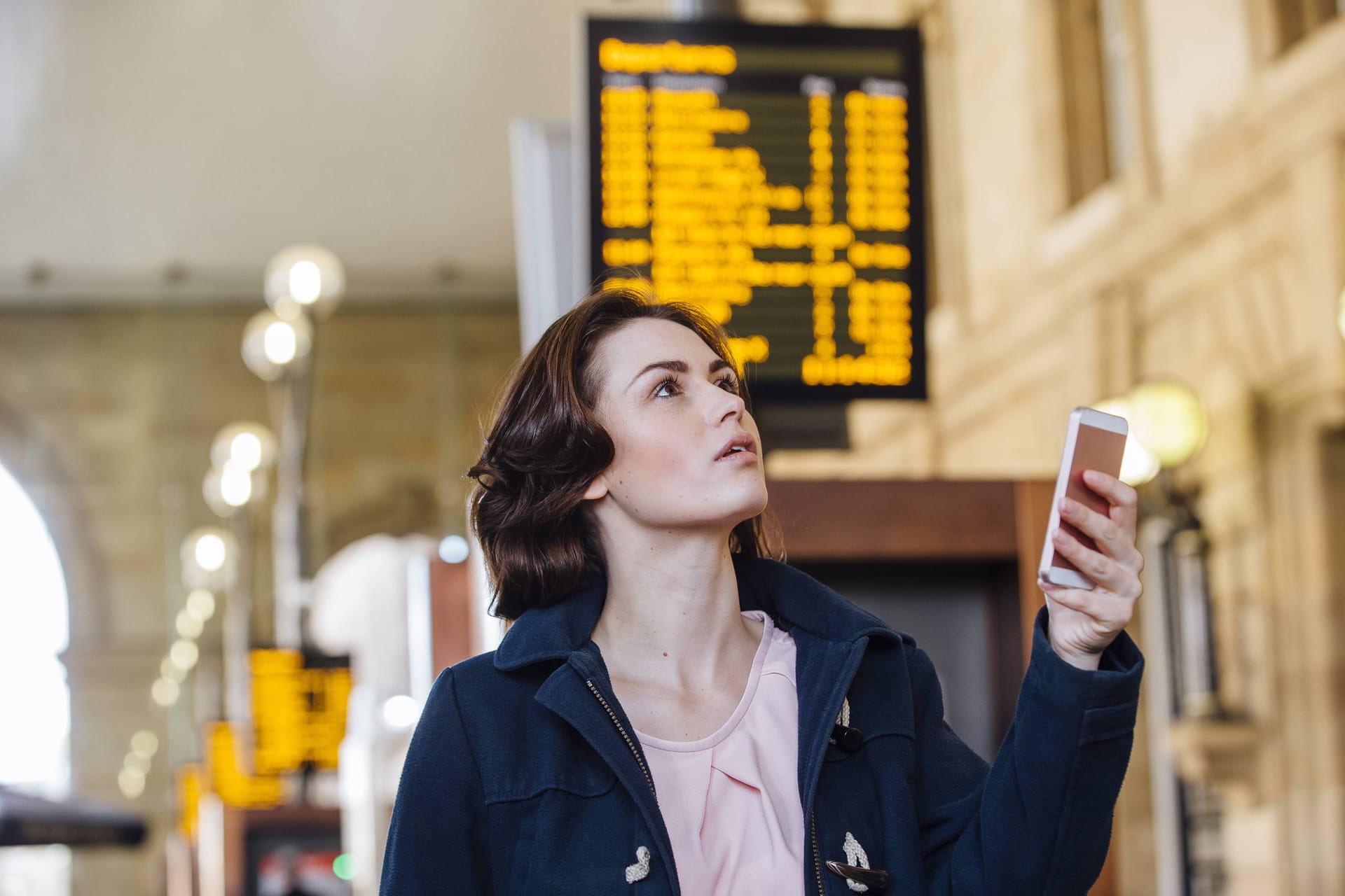 Person checking smartphone while looking up at electronic departure board with yellow text in train station.