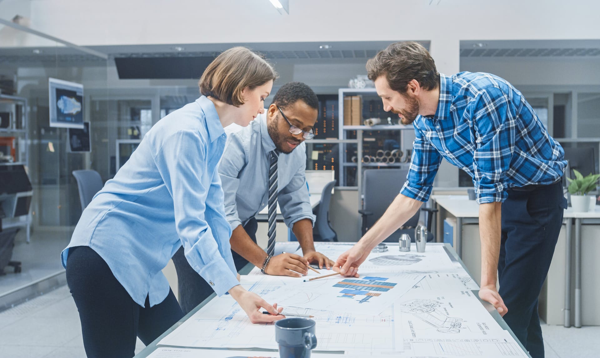 Three colleagues reviewing blueprints and charts on a table in a modern office space with glass partitions.