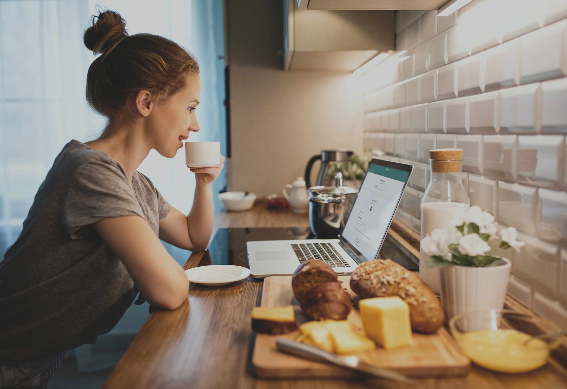 Person drinking coffee while using laptop in kitchen with bread and cheese on wooden board and white tiled backsplash.