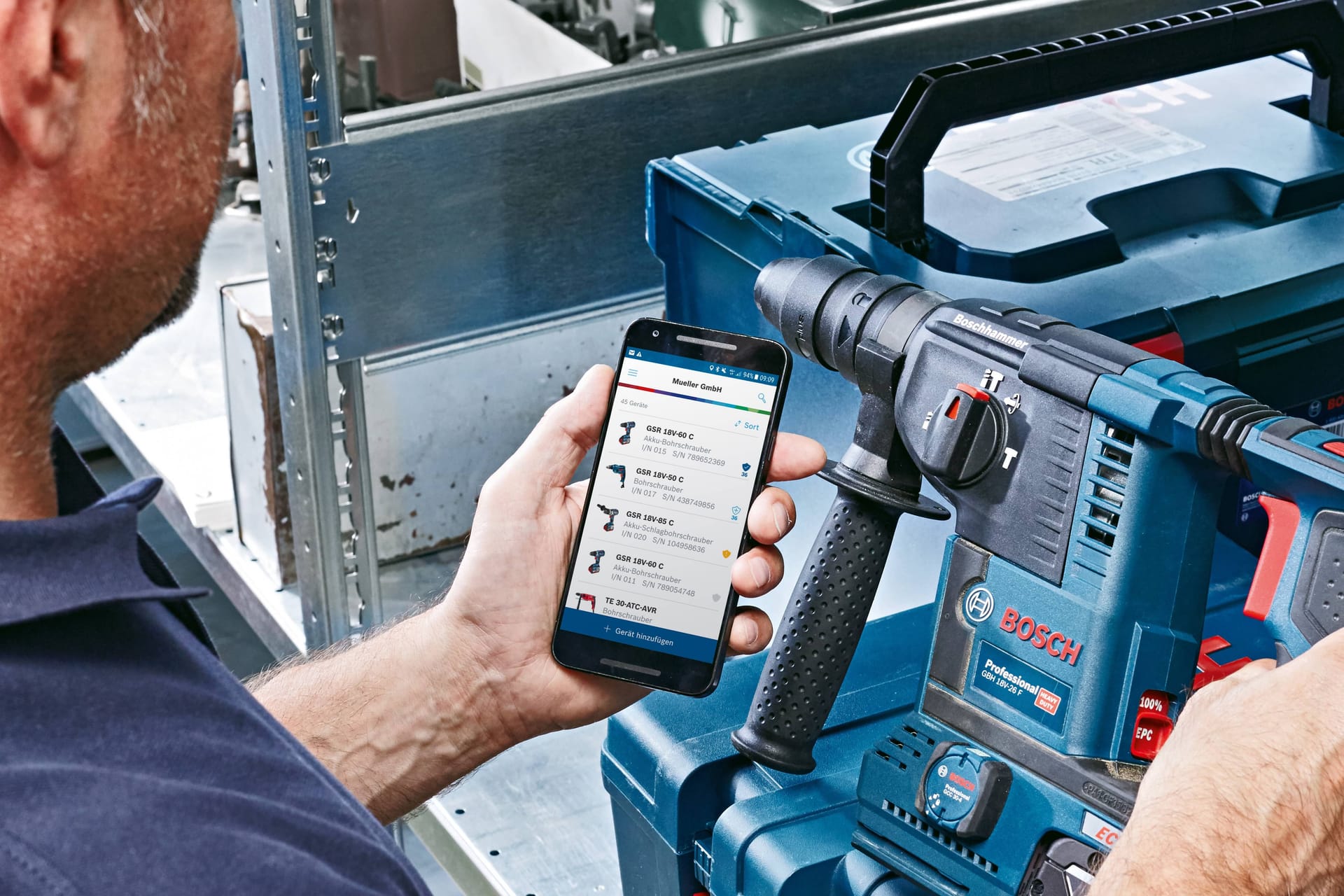 Person checking Bosch power tool inventory on smartphone app next to blue Bosch rotary hammer drill in carrying case.