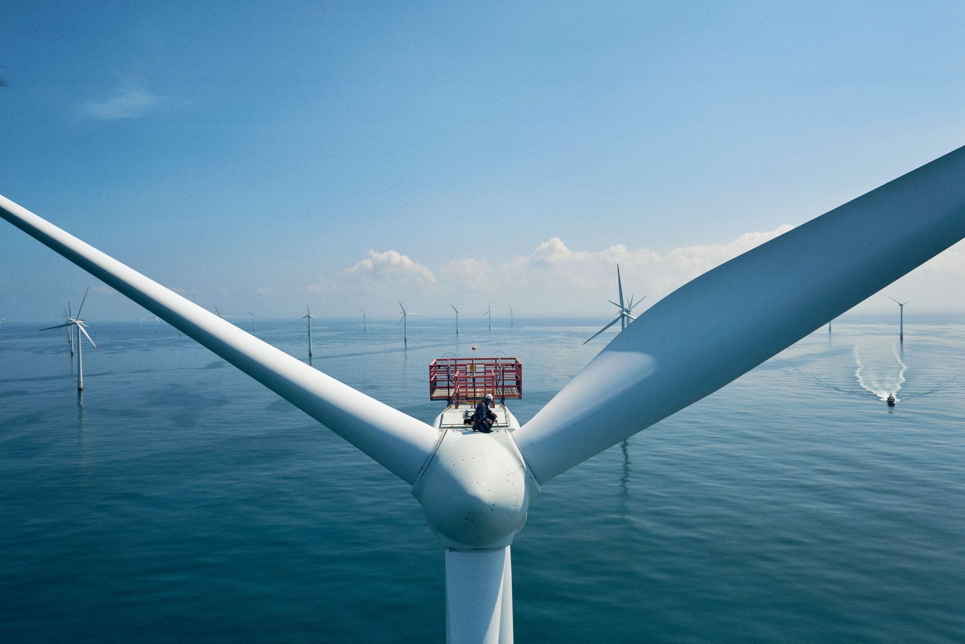 Close-up of offshore wind turbine with maintenance platform, overlooking wind farm on calm blue sea under clear sky.
