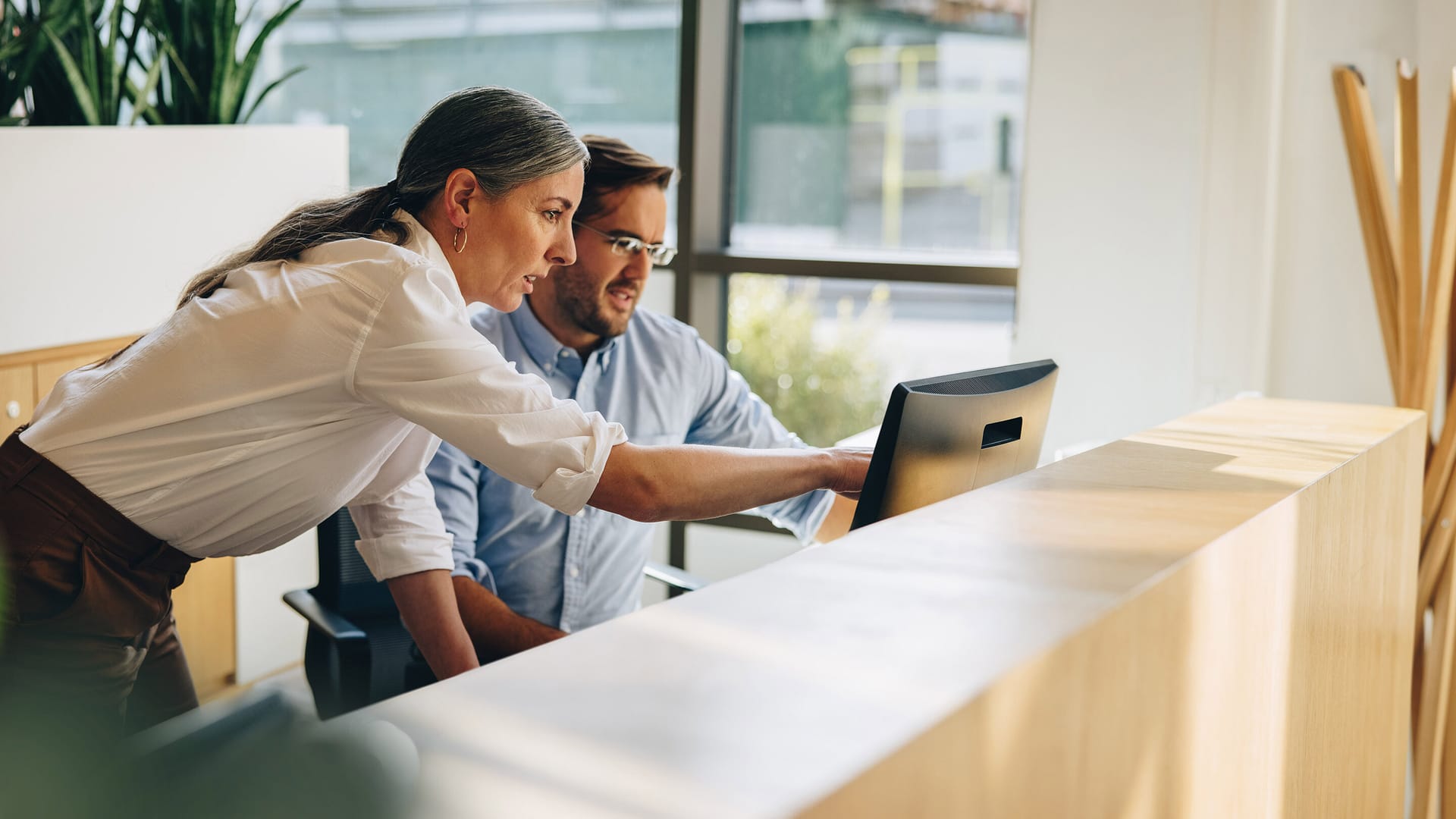 Two colleagues looking at a computer screen at a reception desk in a bright, modern office with plants nearby.