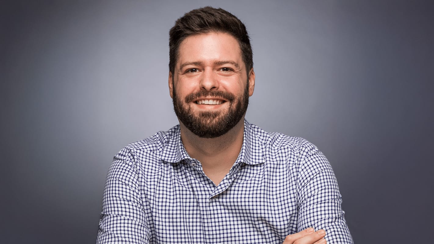 Person with beard smiling against grey background, wearing blue and white checked shirt.