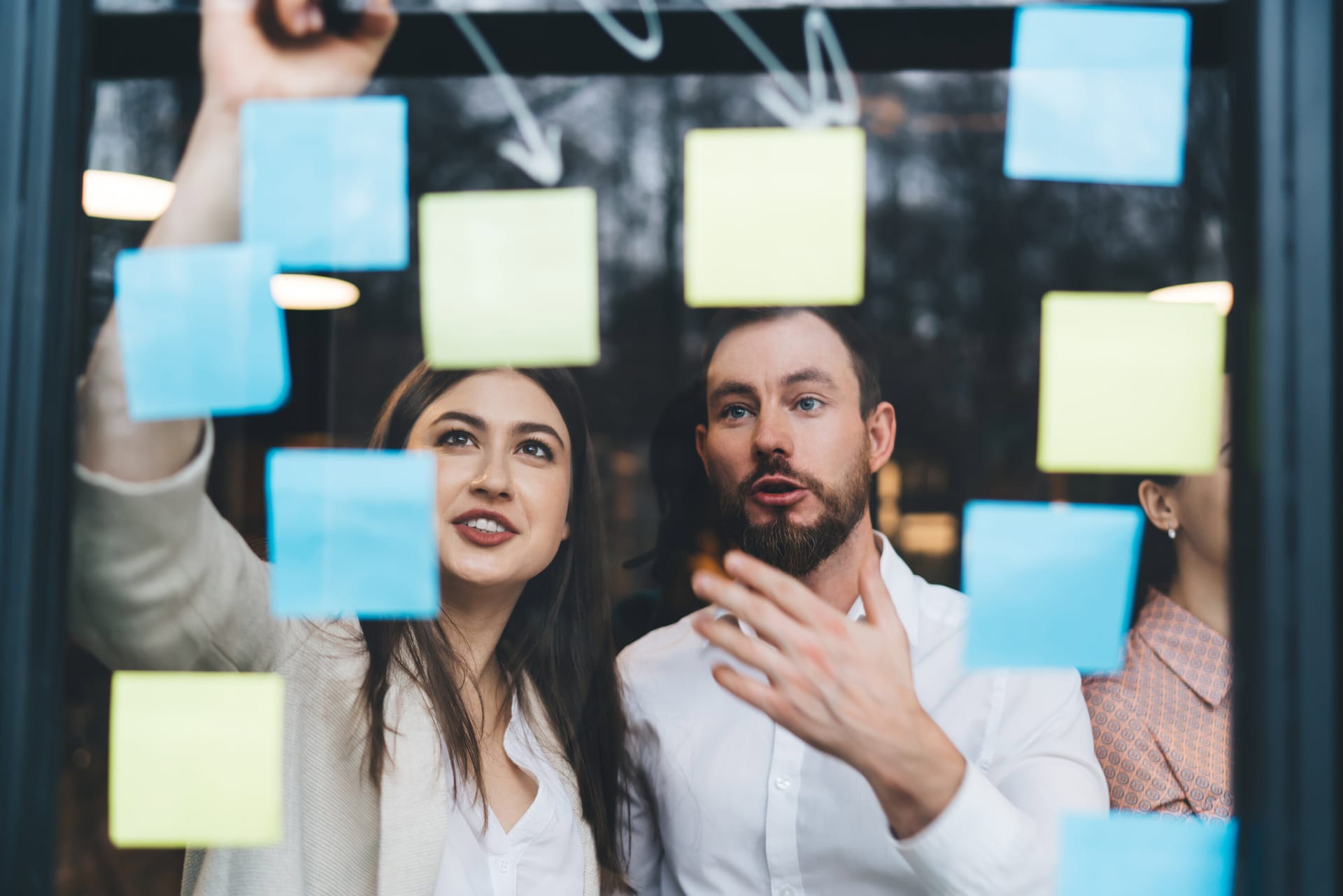 Business colleagues brainstorming with blue and yellow sticky notes on a glass wall during a strategy meeting.