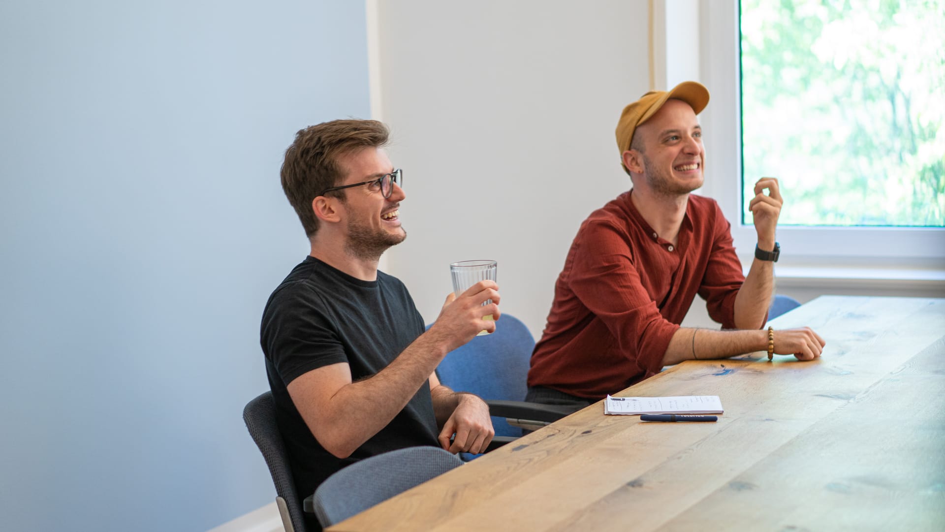 Two smiling men sitting at wooden table in bright office, one in black t-shirt holding drink, other wearing orange cap.