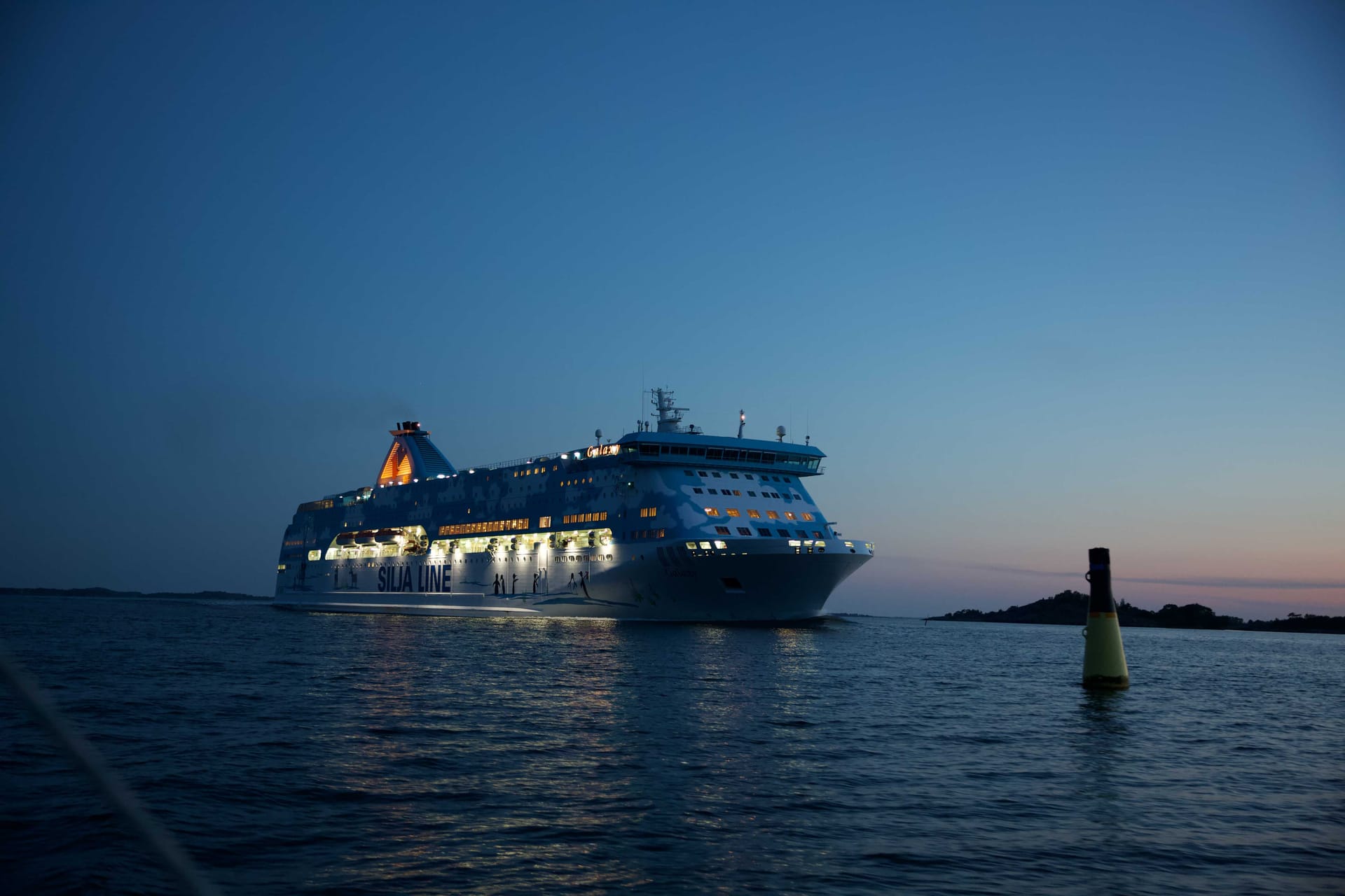 Illuminated Silja Line cruise ferry sailing on calm blue waters at dusk, with navigation buoy visible in foreground.