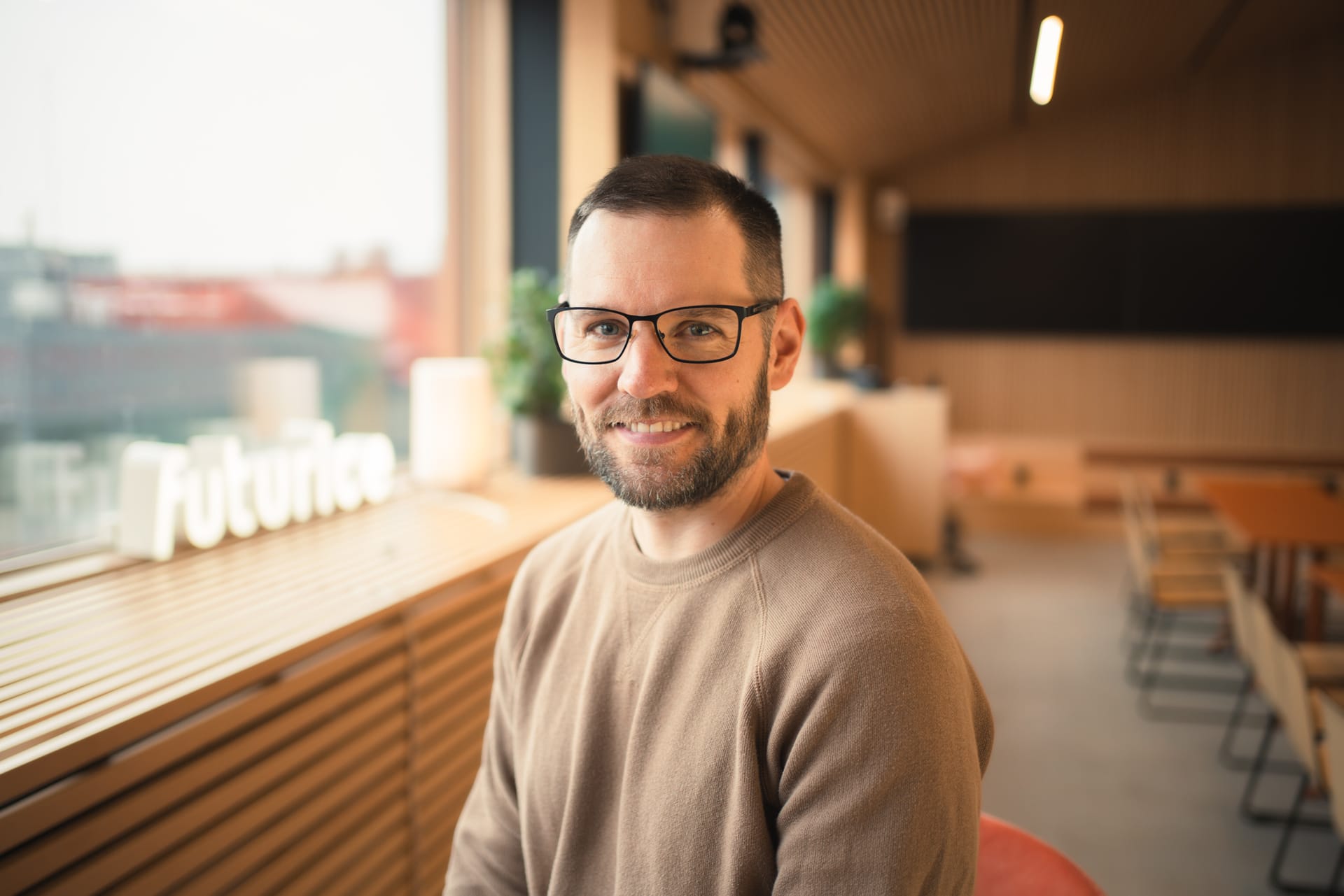 Person smiling in modern office space with wooden interior, large windows, and city view in background.