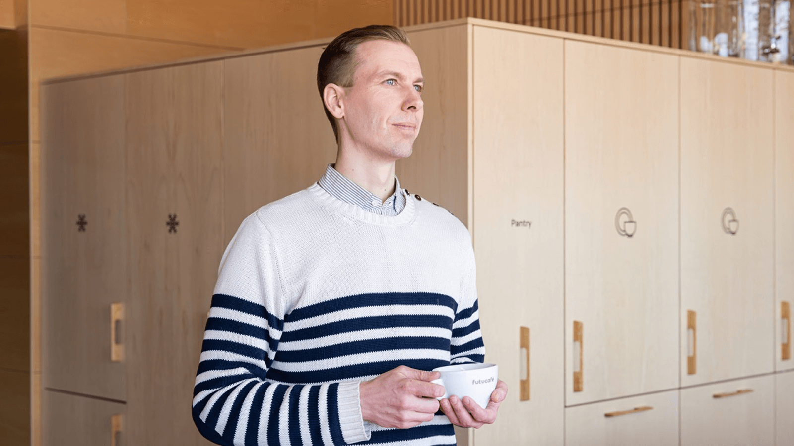 Person in striped navy and white jumper holding a white cup in modern kitchen with wooden cabinetry.