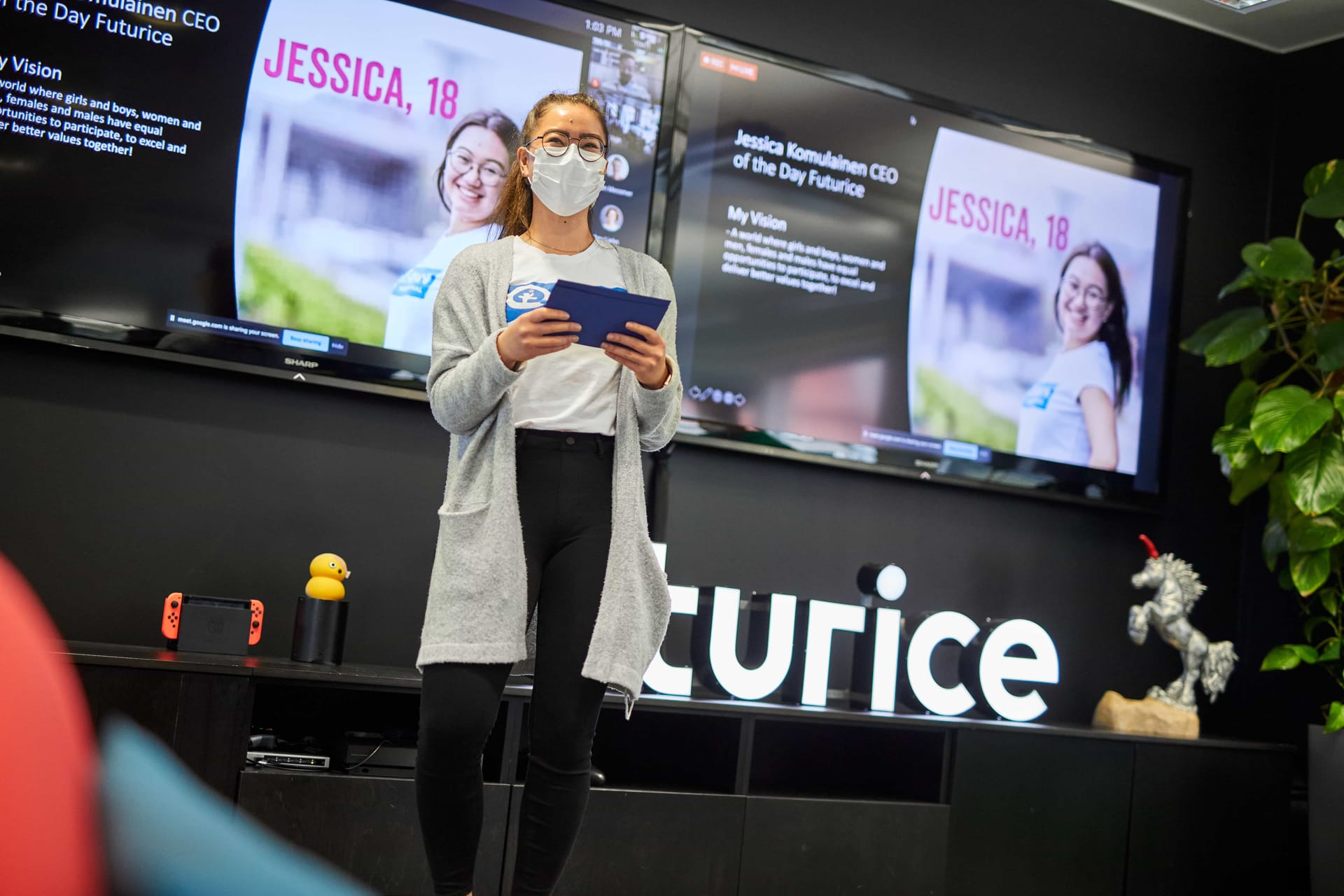 Person wearing mask and grey cardigan presenting at Futurice event, holding blue tablet with screens showing "Jessica, 18" behind.