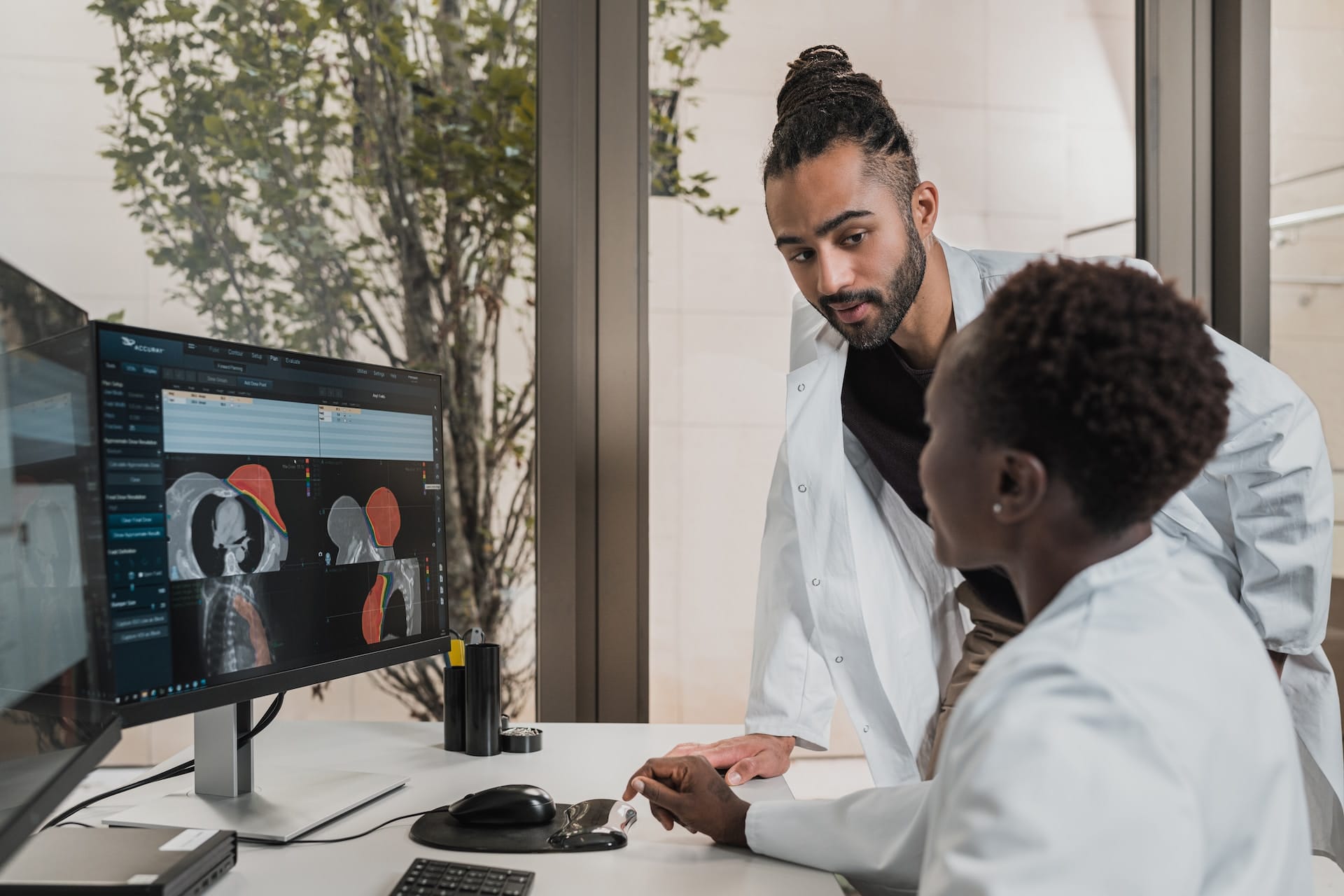Two healthcare professionals in white coats examining medical scans on a computer monitor in a bright office setting.
