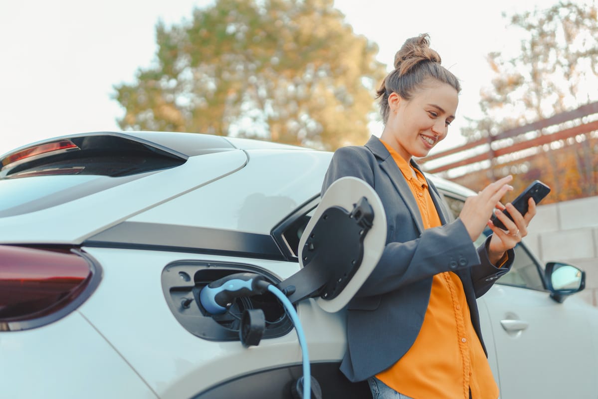 Person in grey blazer and orange shirt using smartphone while charging electric vehicle outdoors in autumn setting.