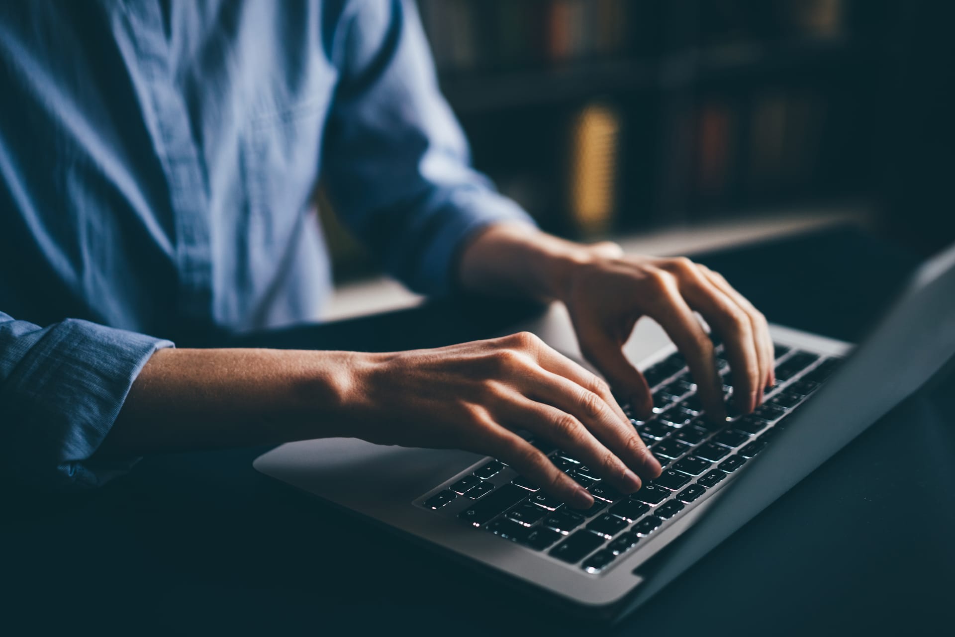 Close-up of hands typing on a laptop keyboard in dim lighting, person wearing a light blue shirt.
