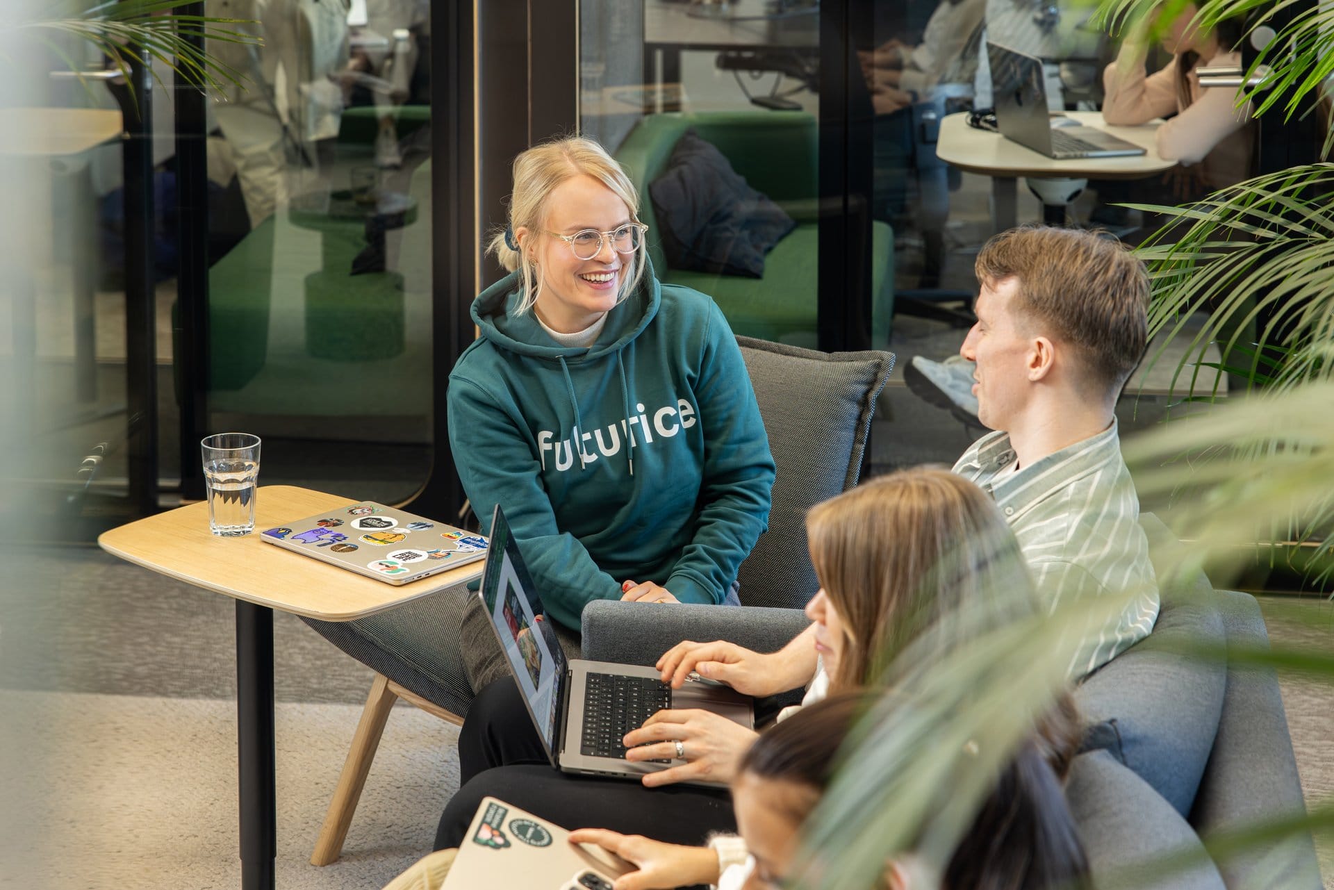 People in a modern workspace having a discussion, with a person in teal "Futurica" hoodie smiling at colleagues with laptops.