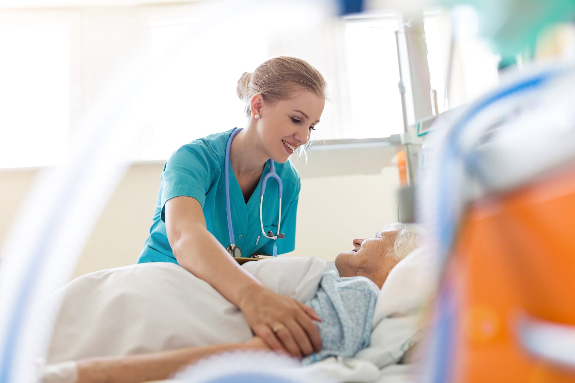 Healthcare worker in teal scrubs with stethoscope attending to elderly patient lying in hospital bed.