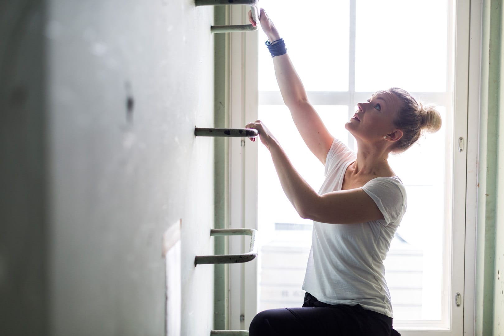 Woman in white top adjusting wall shelves near a bright window, wearing a blue bracelet.