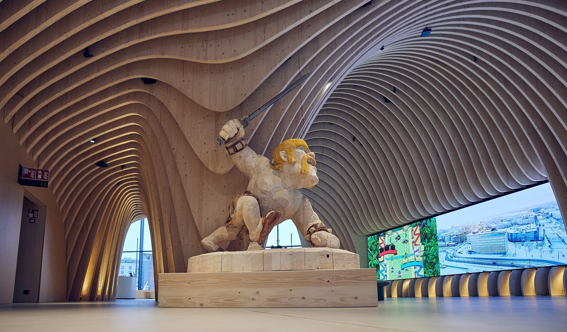 Large sculpture of a figure with golden hair wielding a sword beneath curved wooden ceiling in modern exhibition space.