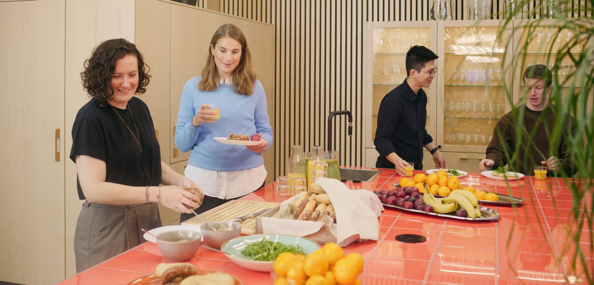 Four people enjoying breakfast at a bright orange table with fresh fruit, bread and juice in a modern kitchen space.