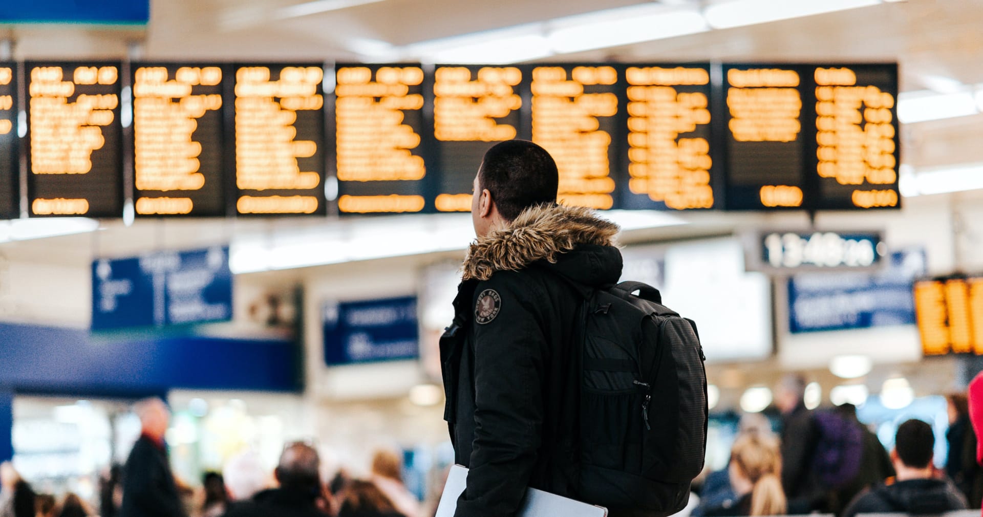 Traveller in winter coat with backpack looking at orange departure boards in busy train station terminal.