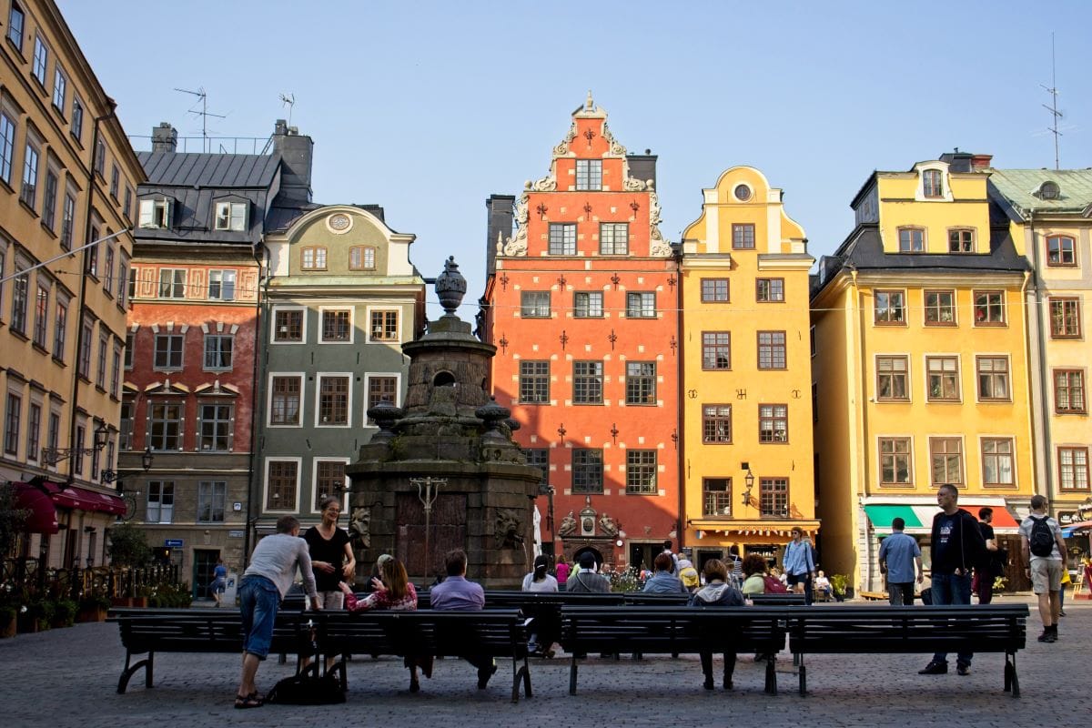 Colourful historic buildings in Stockholm's Gamla Stan square with people sitting on benches and a stone fountain in the foreground.